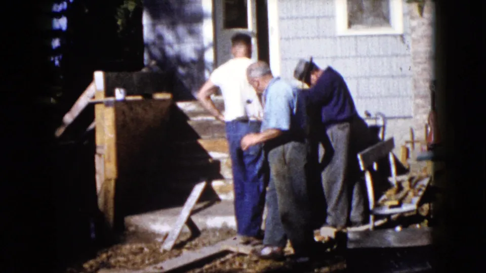 1960 Wauconda frame showing three men gathered beside a wooden structure, hand pump, house siding, and yard steps.