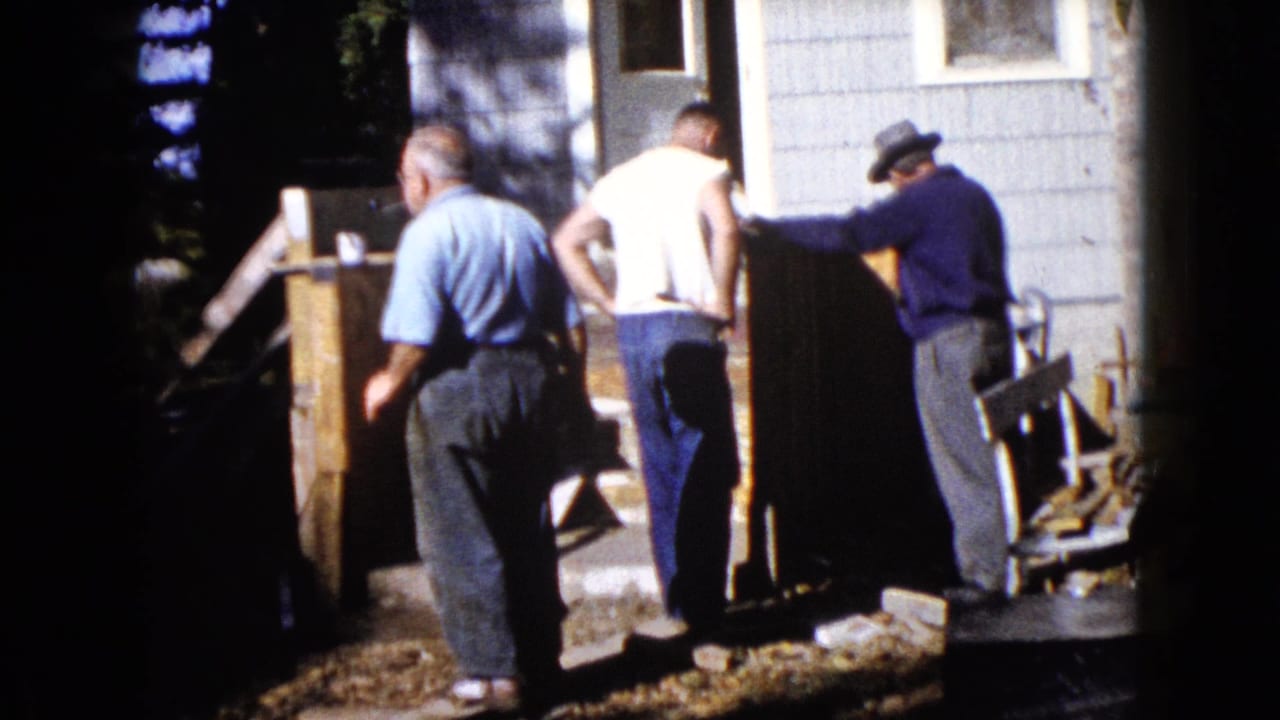 A 1960 home-movie frame shows men near wooden boards and a small entry area outside a Wauconda house.