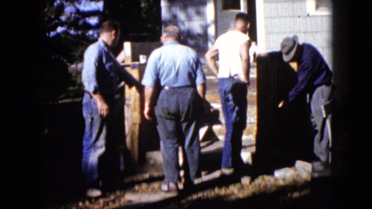 Men shift position around a large wooden item beside house steps.