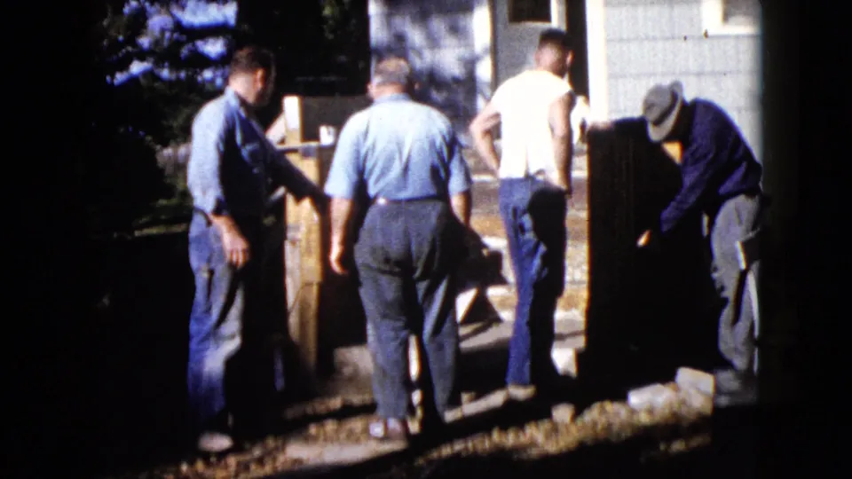 1960 Wauconda frame with several men in work clothes positioning a heavy wooden item near a house entrance.