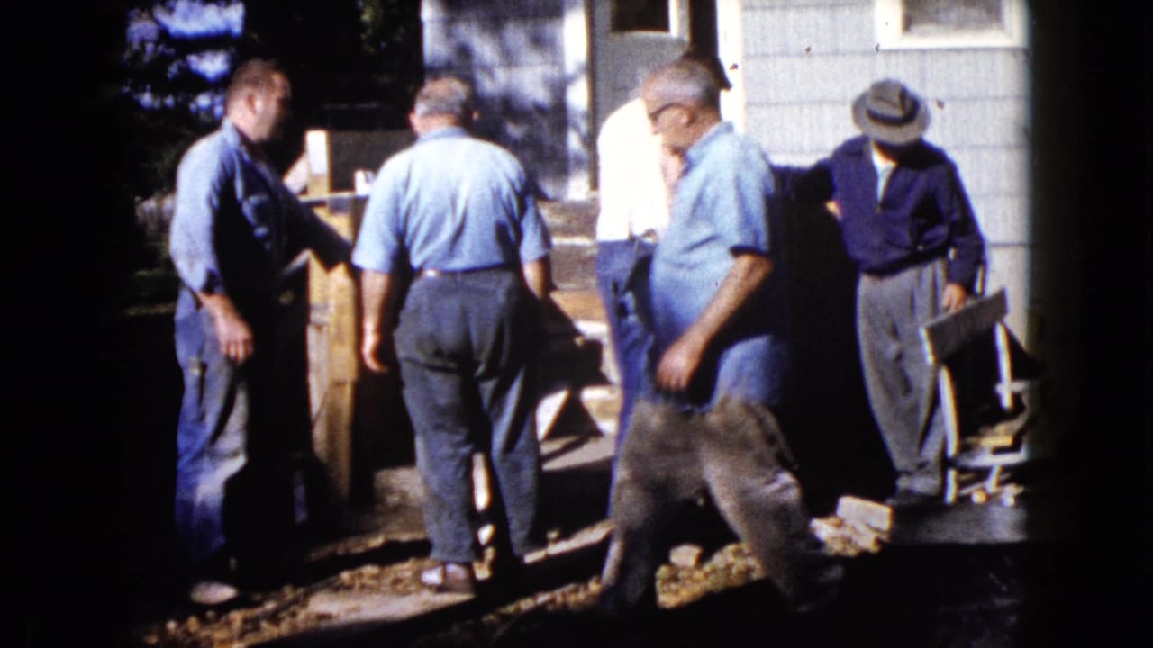 1960 Wauconda home-movie frame showing one man crossing foreground as others work near a wooden object by the steps.