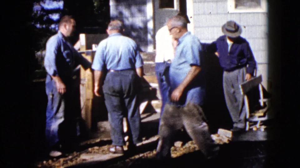 1960 Wauconda home-movie frame showing one man crossing foreground as others work near a wooden object by the steps.
