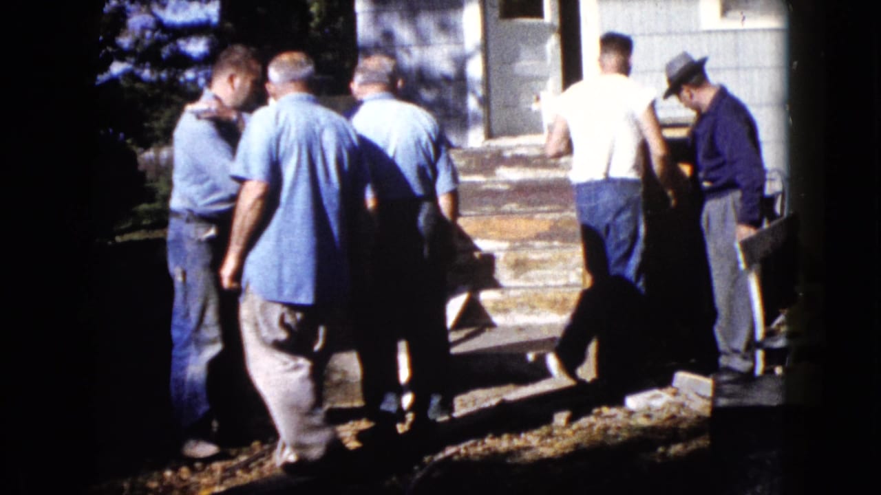 1960 Wauconda frame showing men clustered near a house entrance with a wooden piece and tools or boards nearby.