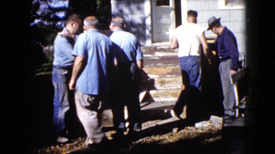 1960 Wauconda frame showing men clustered near a house entrance with a wooden piece and tools or boards nearby.