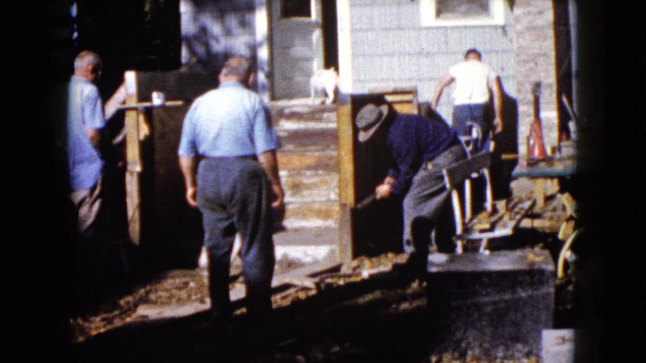 Men work near porch posts while a small white dog stands by the doorway.