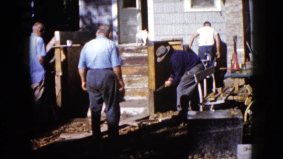 A small white dog stands near the door as men continue work on the wooden porch structure in Wauconda, Illinois.