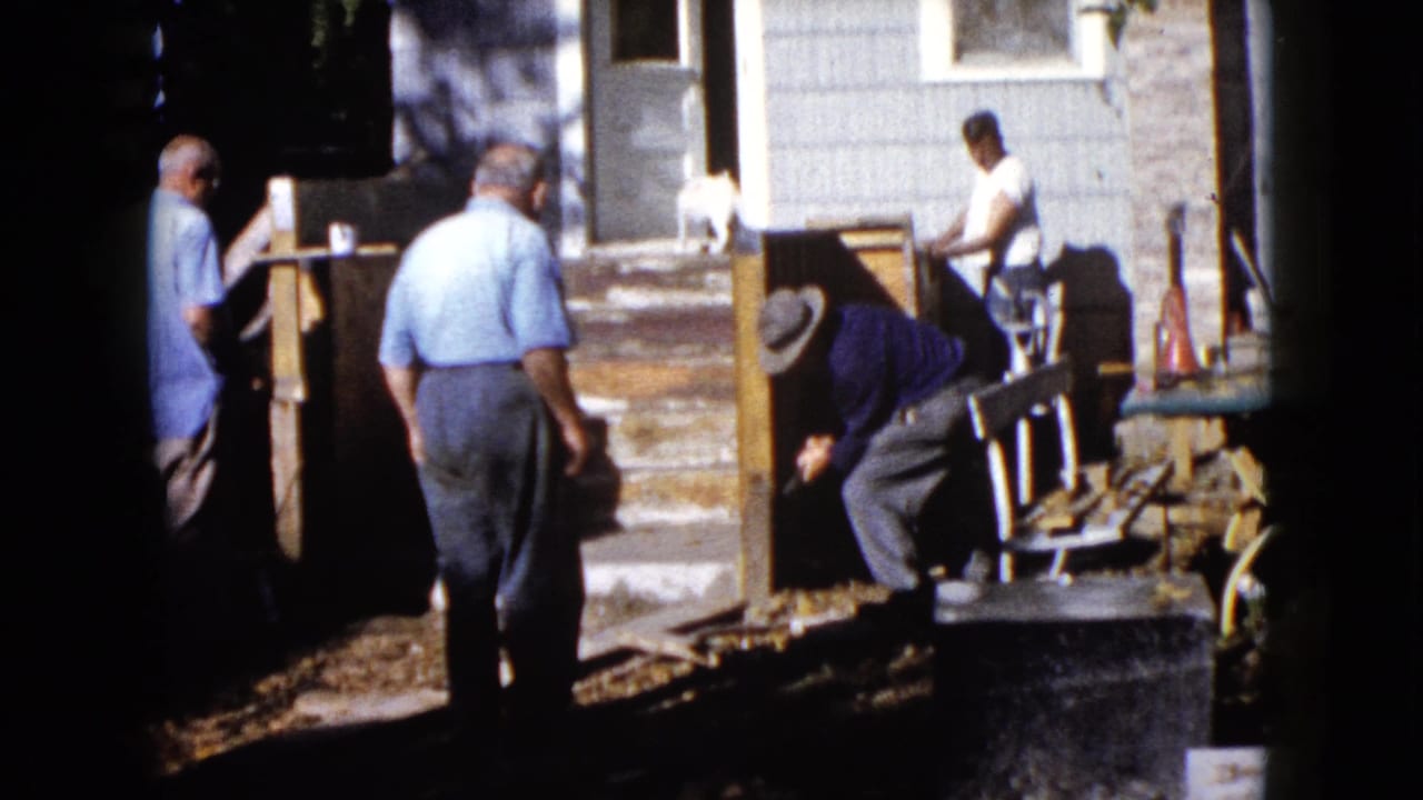 In the 1960 Wauconda home-movie scene, one man bends over porch materials while others stand nearby in the yard.