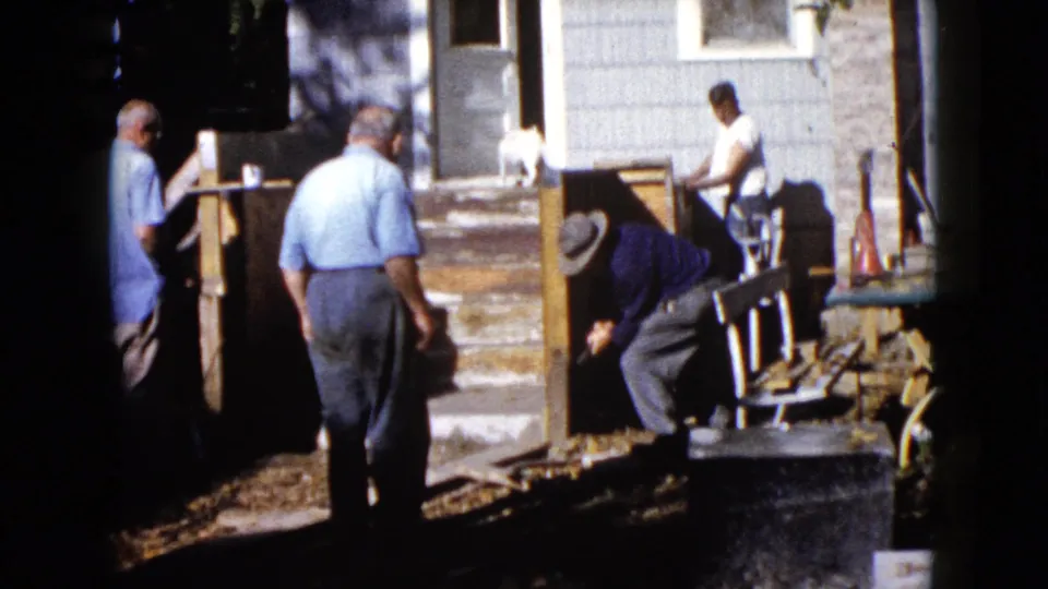 In the 1960 Wauconda home-movie scene, one man bends over porch materials while others stand nearby in the yard.