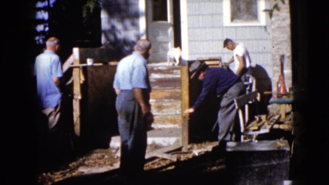The porch work continues beside the house, with men positioned around the steps and one man bent over the wooden structure.