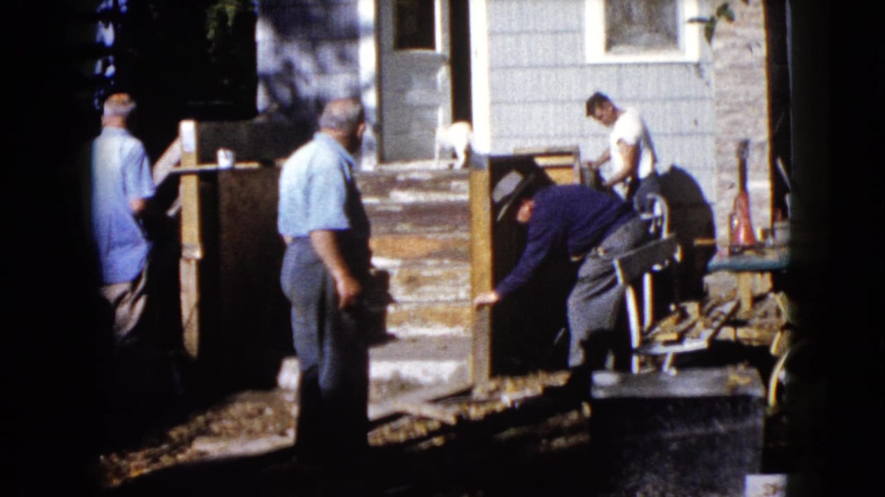 Men working outside a house with porch steps, boards, and a small white dog nearby.