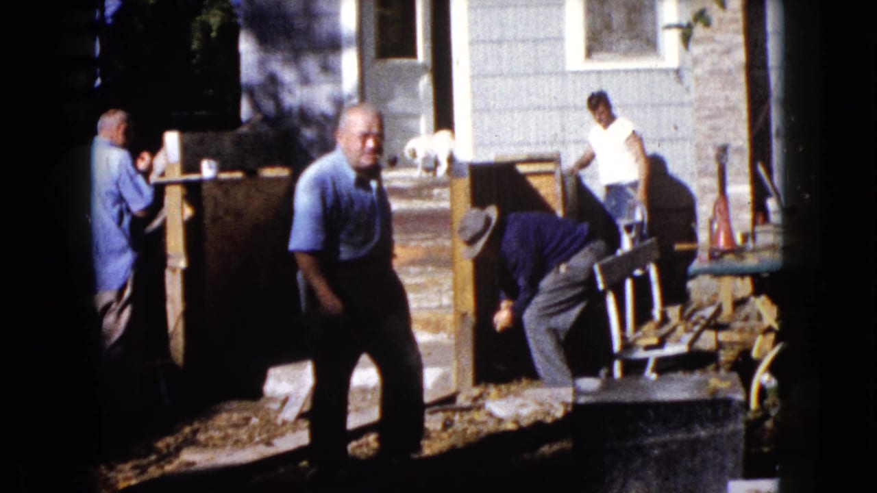 1960 Wauconda frame with one man approaching the camera as two others continue work near a house porch and wooden materials.