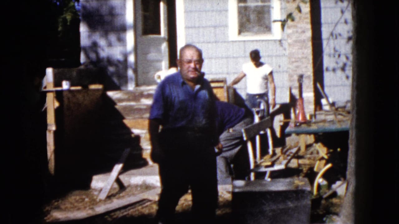1960 Wauconda home-movie frame showing a man near the camera, another man by the porch, and repair materials beside a house exterior.