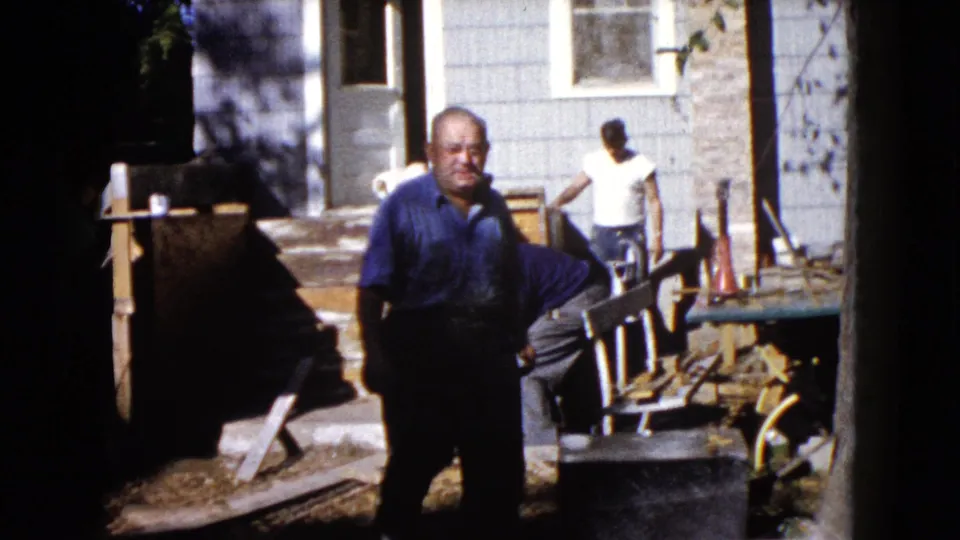 1960 Wauconda home-movie frame showing a man near the camera, another man by the porch, and repair materials beside a house exterior.