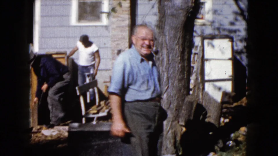 1960 Wauconda frame with a smiling man near a tree trunk and other men working around porch materials at a residential exterior.