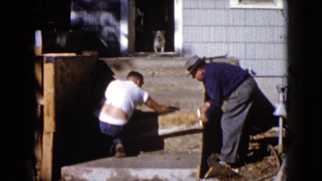 Two men work on wooden exterior steps while a dog sits in the doorway behind them.