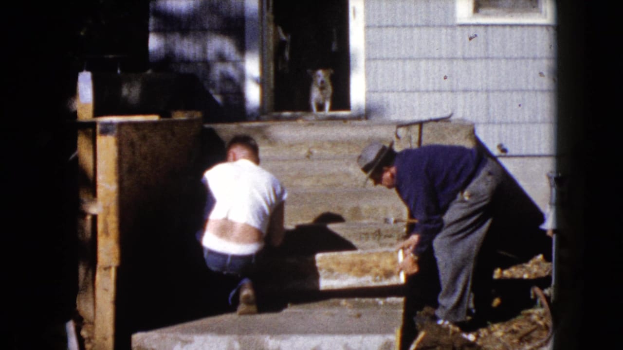 A 1960 Wauconda home-movie frame shows two men working at a wooden exterior stairway beside a light-sided house.