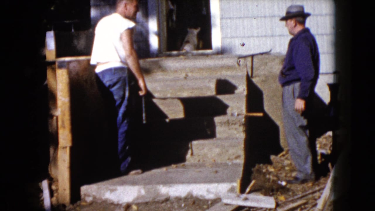 Two men stand outside a house near a concrete porch, with a cat visible in the doorway.