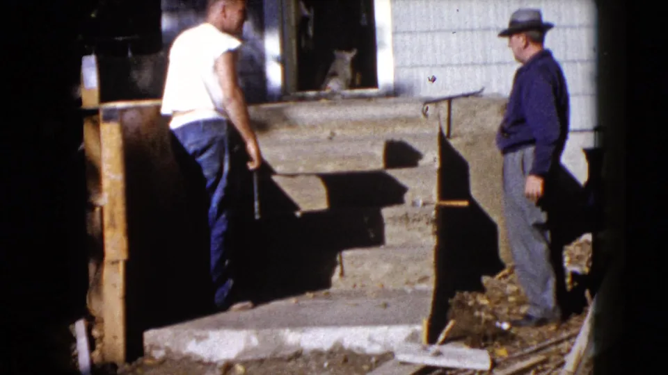 In Wauconda, Illinois, two adult men stand near a porch and house exterior during a 1960 home-repair scene, with a cat in the doorway.