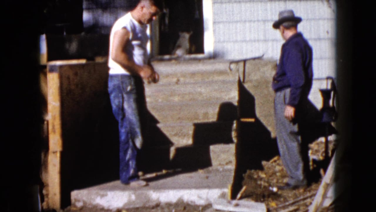 A 1960 Wauconda home-movie frame shows one man by the porch and another in a fedora standing near the house entrance.