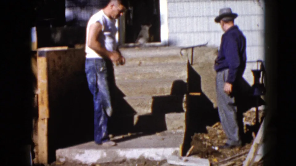 A 1960 Wauconda home-movie frame shows one man by the porch and another in a fedora standing near the house entrance.