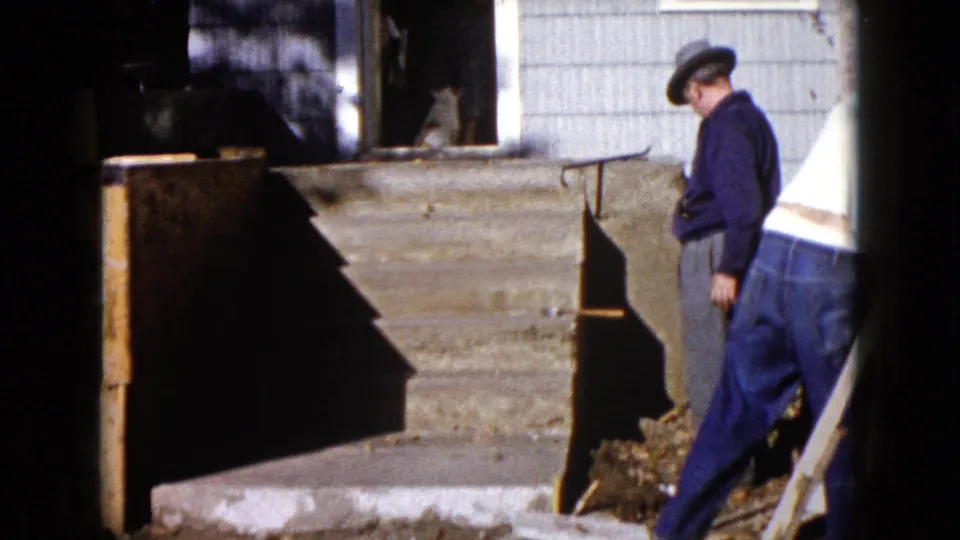 At the end of the clip, a fedora-wearing man stands by the house exterior while the other man moves out of frame.