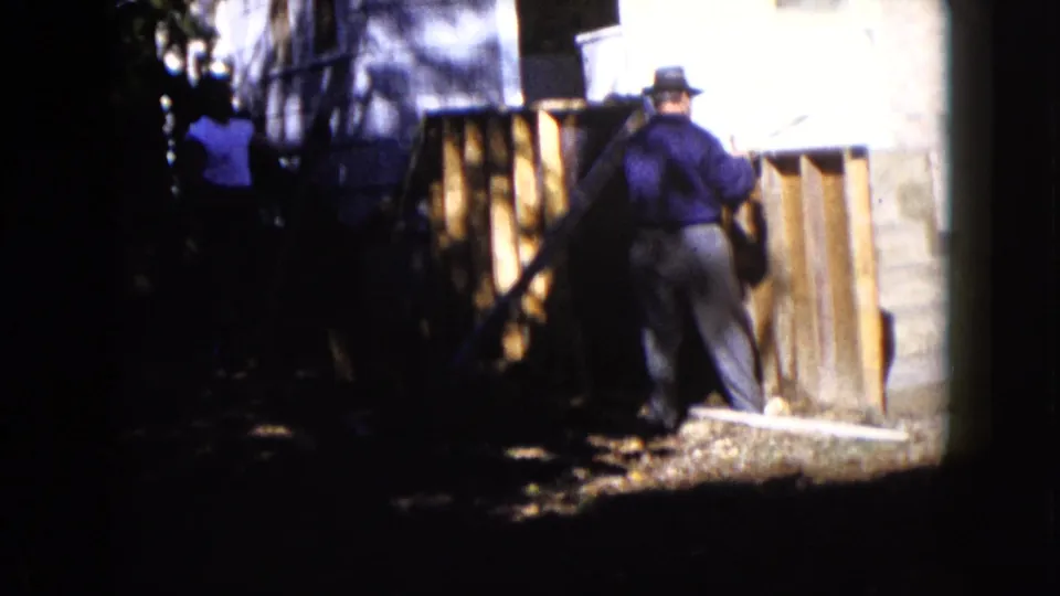 Two adult men are visible near a house in 1960 Wauconda footage, with one working at vertical boards by concrete steps.