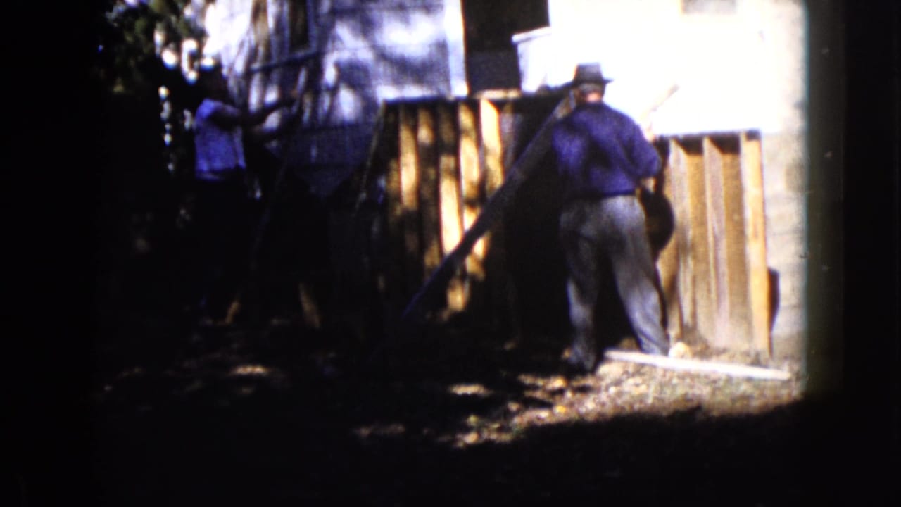 Men stand near a wooden structure beside a light-sided house in deep shade and sun.