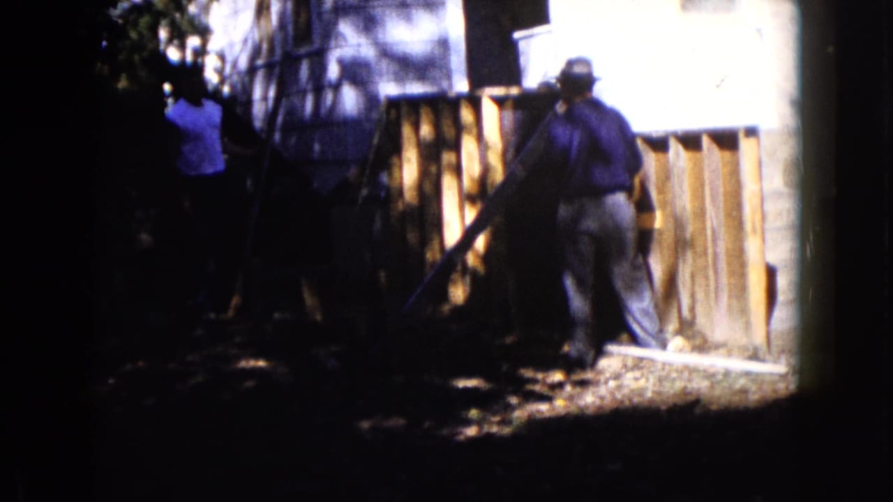 A hat-wearing man works beside a wooden structure outside a house in the Wauconda home-movie scene.