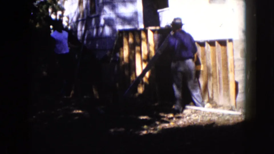 A hat-wearing man works beside a wooden structure outside a house in the Wauconda home-movie scene.