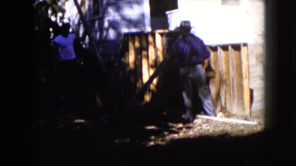 The 1960 Wauconda home movie shows men clustered around boards and a wooden structure beside a house.
