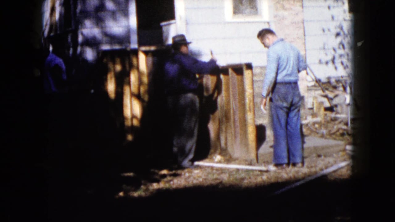 Two men stand beside upright wooden boards near a house exterior.