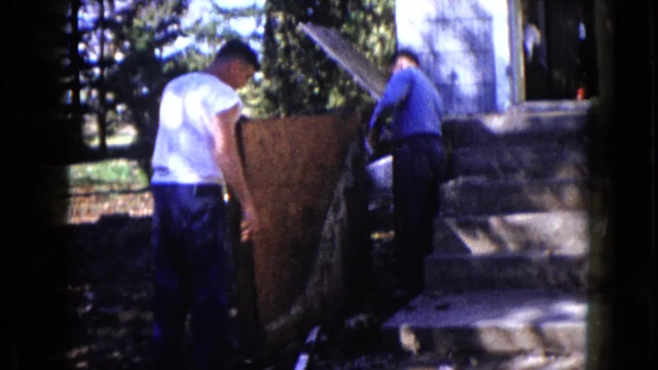 A man in a white shirt and another in blue work near a large wood panel by house steps in 1960 Wauconda footage.