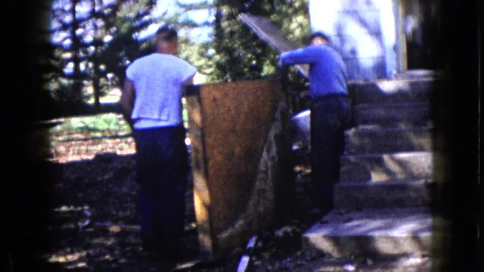 Two adult men work with a large wood panel beside steps and trees in Wauconda, Illinois archival home-movie footage.