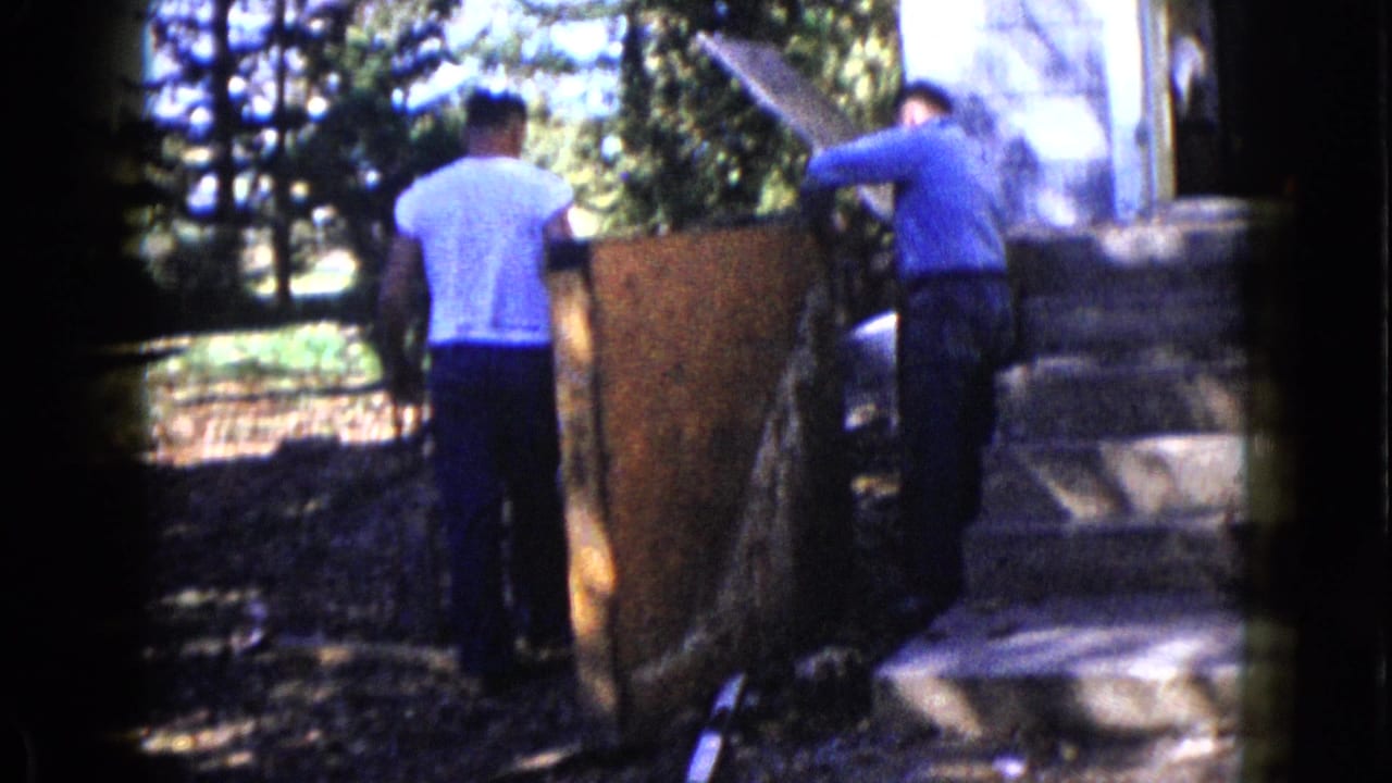 In 1960 Wauconda home-movie footage, two men work with a large wood panel near a house entrance shaded by trees.