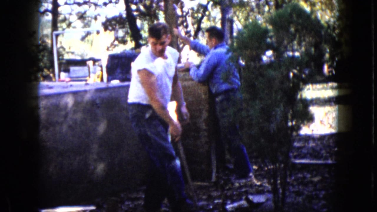 Two men work outdoors near a low wall, one holding a tool in the foreground.