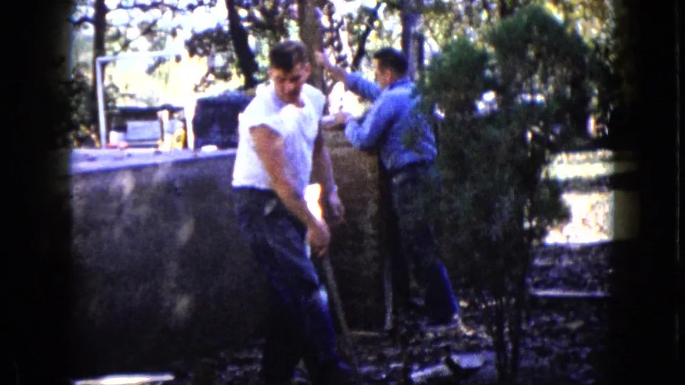 In 1960 Wauconda, Illinois, two adult men work in a shaded yard near a low concrete wall and trees.