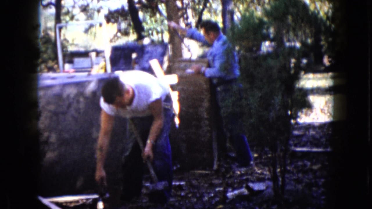 One man in a white T-shirt bends over a tool while another works near the wall in the 1960 Wauconda yard scene.