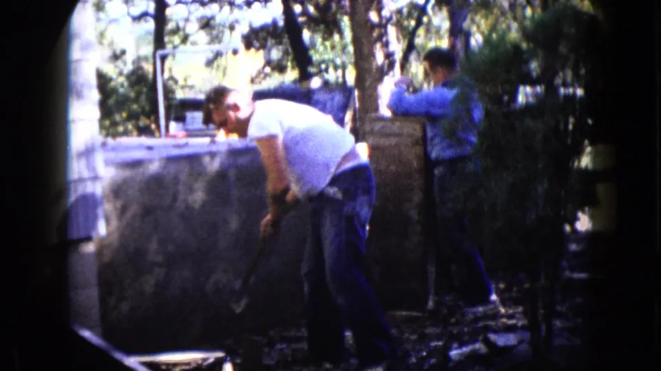 In the final frame, a man in a white T-shirt works beside the wall as another man stands deeper in the yard.