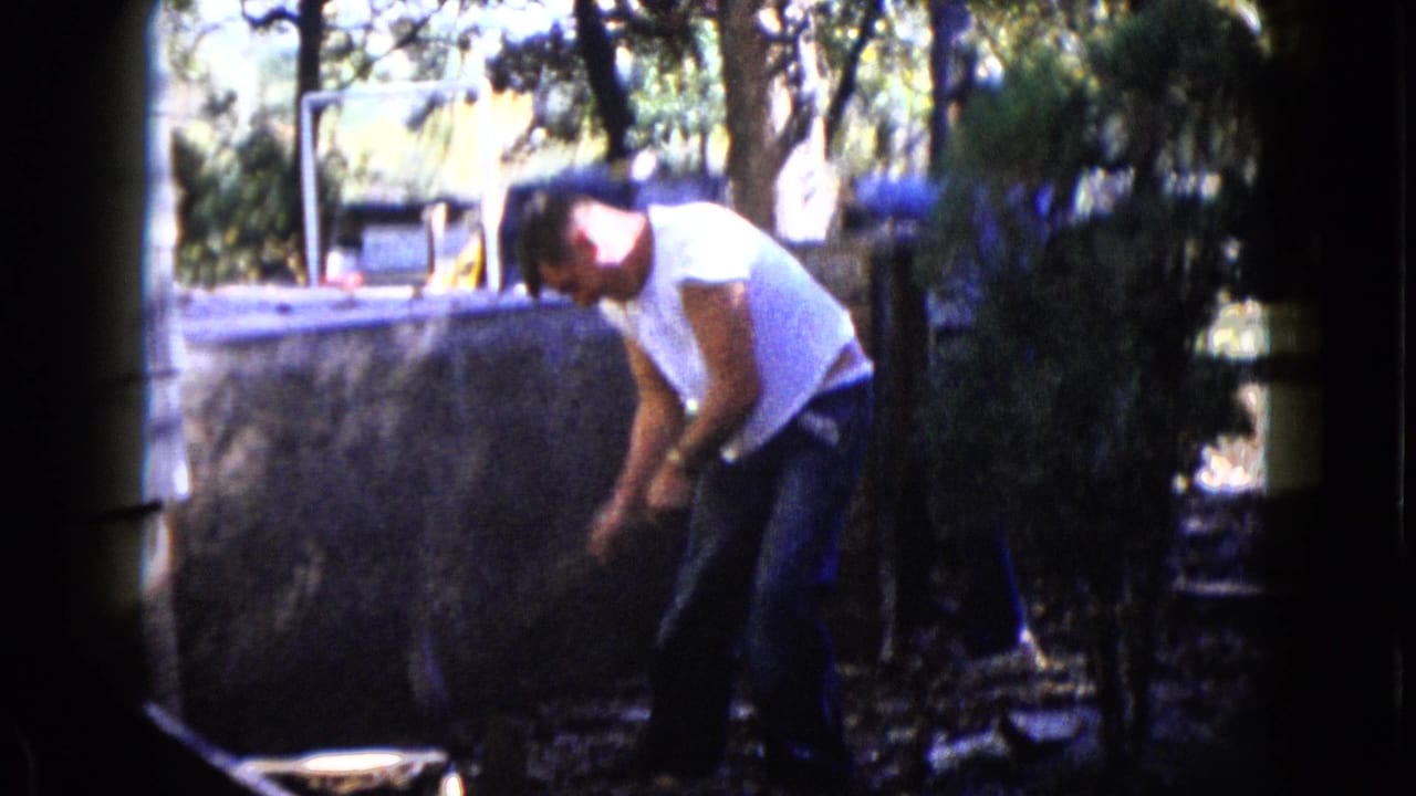 Man in a white T-shirt bends over a backyard work area with a sledgehammer near a low wall.