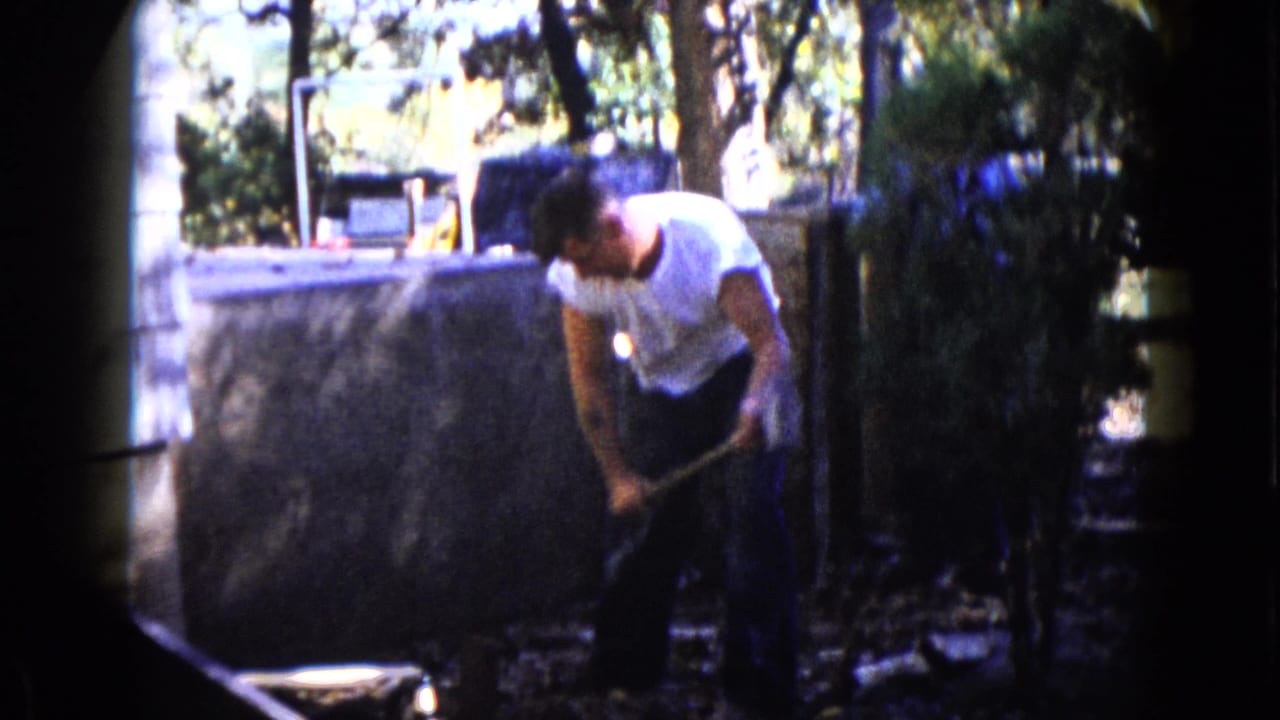 The man continues hammering or repositioning the sledgehammer in a shaded Wauconda backyard, with trees and a low wall behind him.