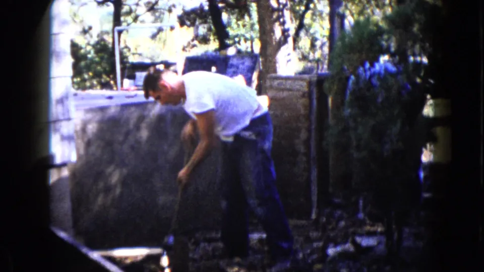 A man works low to the ground with a sledgehammer as another person in blue appears nearby in the 1960 Wauconda backyard scene.
