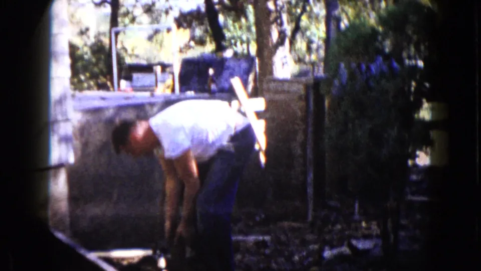 The man bends over the backyard work area with the sledgehammer near the ground, framed by trees, a wall, and strong 8mm color contrast.