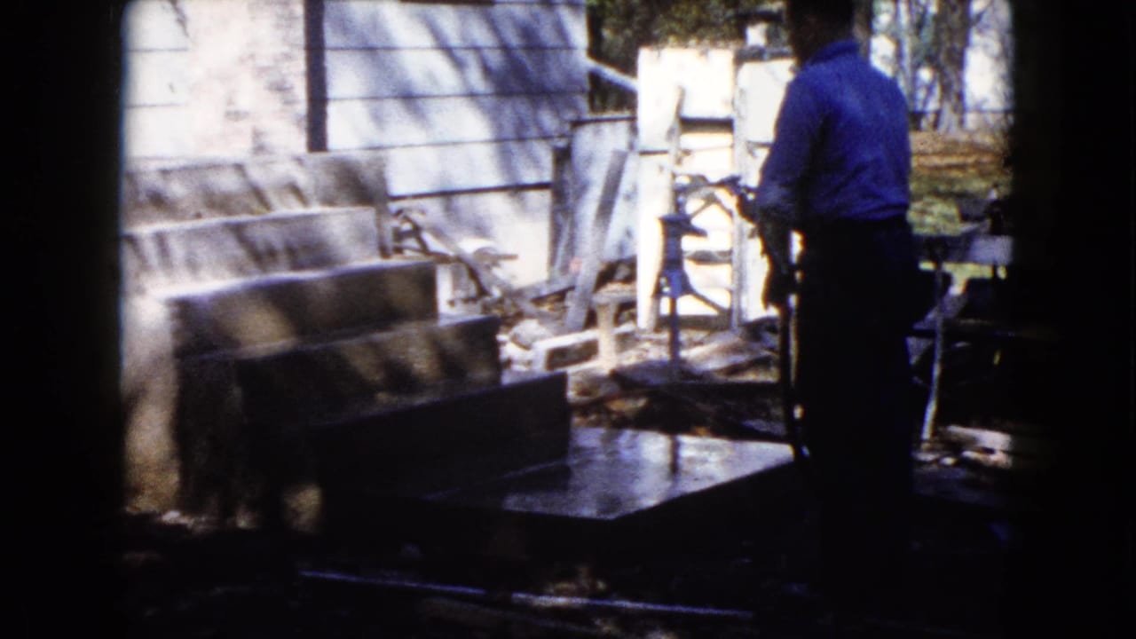 A 1960 Wauconda home-movie frame shows a man beside exterior concrete steps with water and yard equipment nearby.