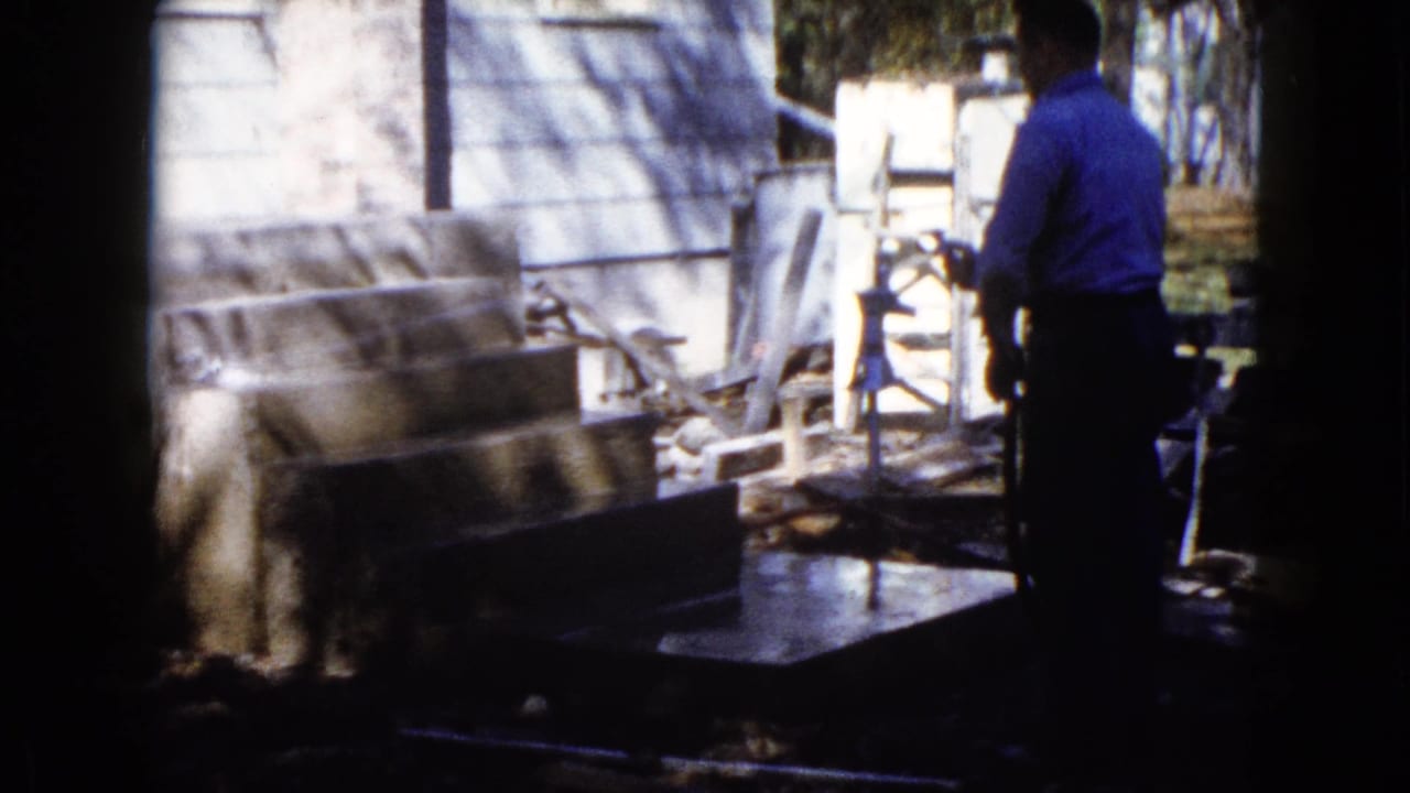 The man continues watering the concrete area beside a light house exterior in the 1960 Wauconda home-movie scene.