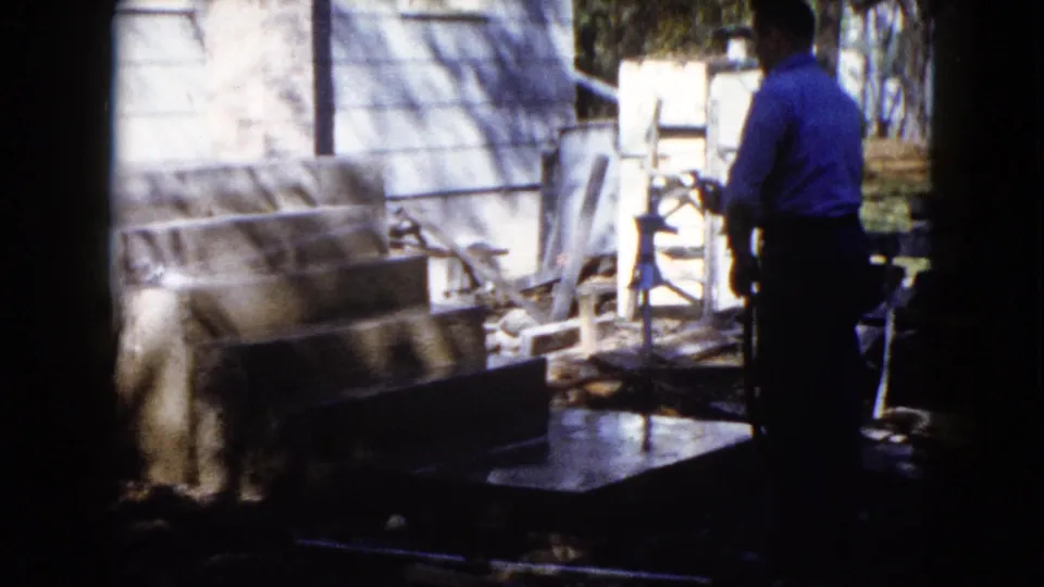 The man continues watering the concrete area beside a light house exterior in the 1960 Wauconda home-movie scene.