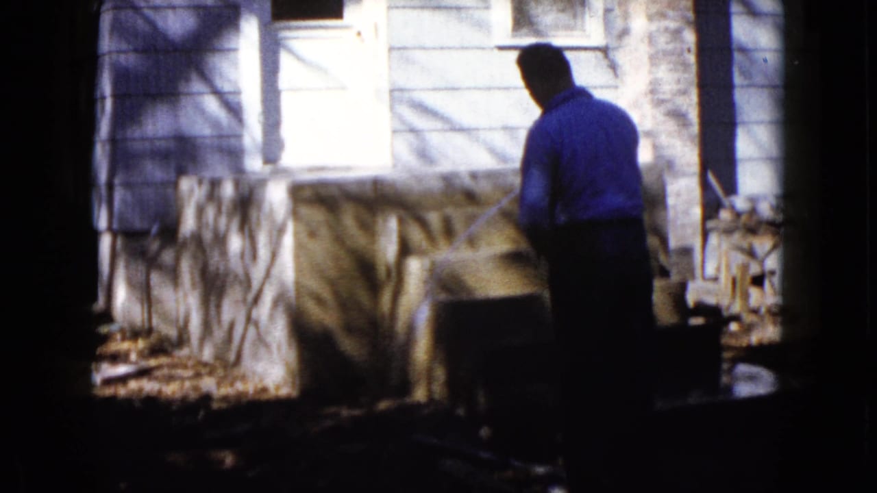 The 1960 Wauconda home-movie frame shows a man from behind watering new concrete steps in bright sunlight beside a house.