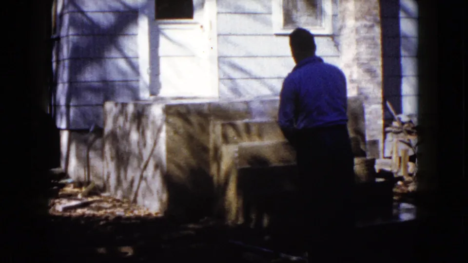 A man in a blue shirt waters the fresh concrete steps of a Wauconda house, with siding, a door, and branch shadows visible.