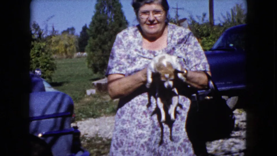 In Wauconda, Illinois, 1960, a smiling woman in a floral dress holds a small dog near a lawn, shrubs, and blue car.