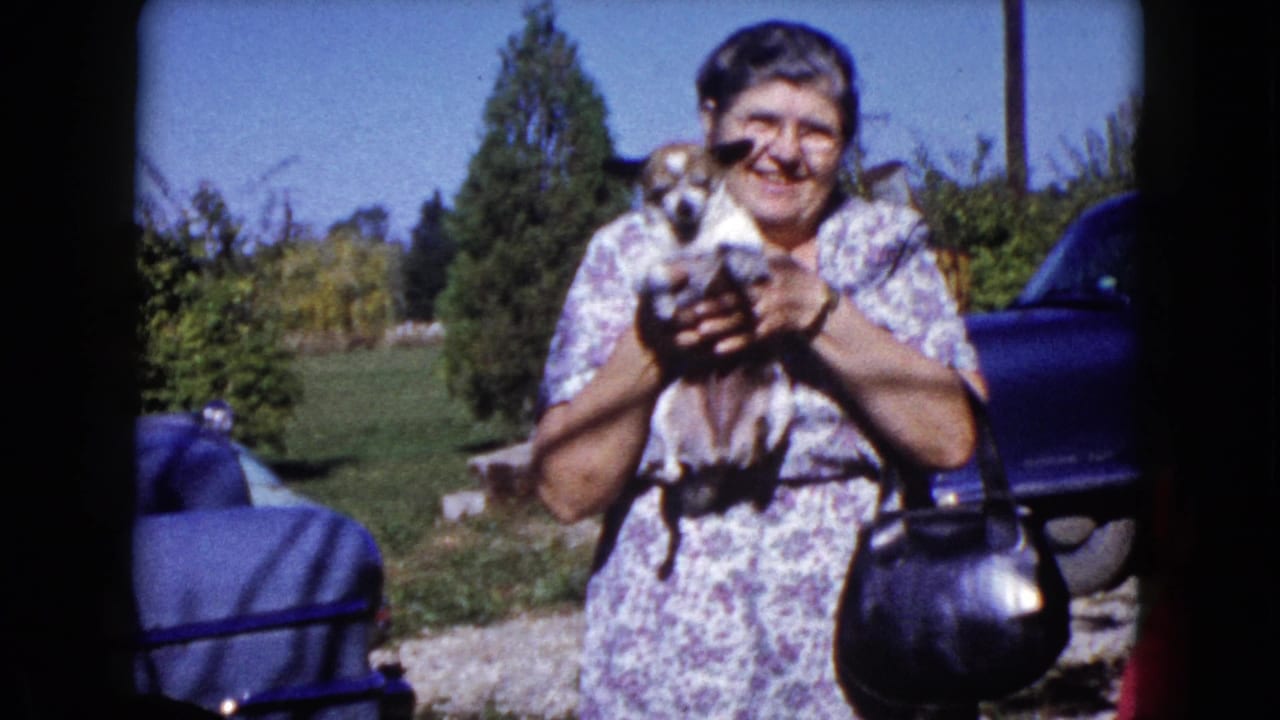 The woman smiles toward the camera as she lifts the small dog in a 1960 Wauconda yard with greenery and a car behind her.