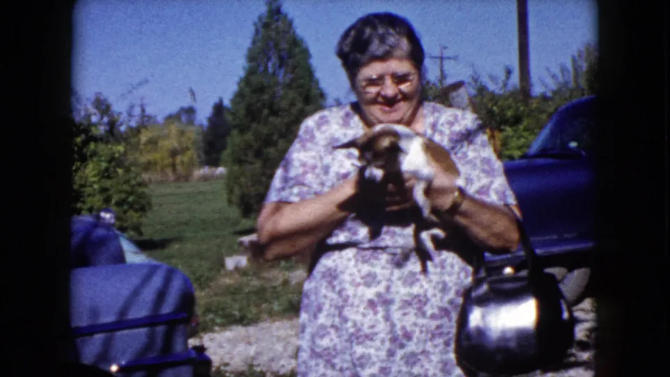 A woman in a floral dress cuddles the small dog close while standing in a Wauconda, Illinois yard in 1960.